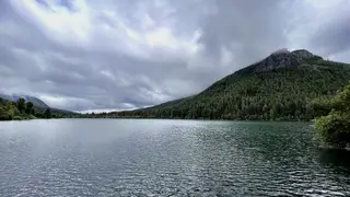 A calm lake stretches beneath a cloudy sky, bordered by dense forest and a steep rocky mountain.