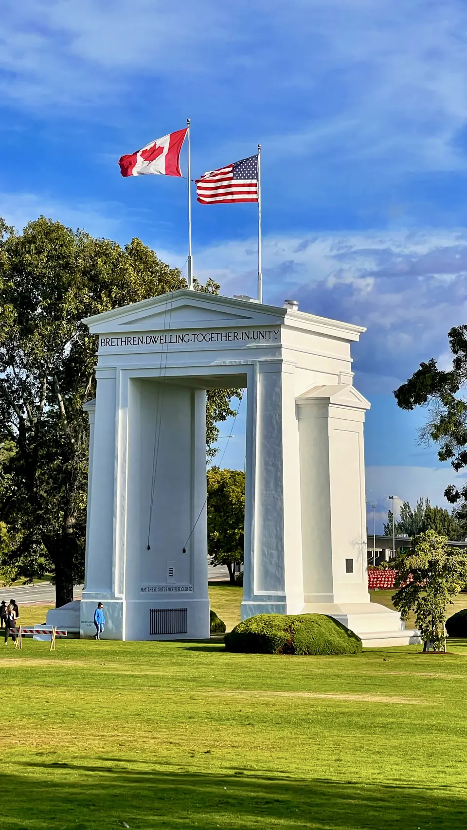 A white memorial arch inscribed “BRETHREN, DWELLING, TOGETHER, IN UNITY” stands on a grassy lawn beneath Canadian and American flags.