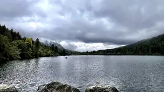 A rippling lake stretches between forested hills beneath a low blanket of gray clouds, with small boats on the water.