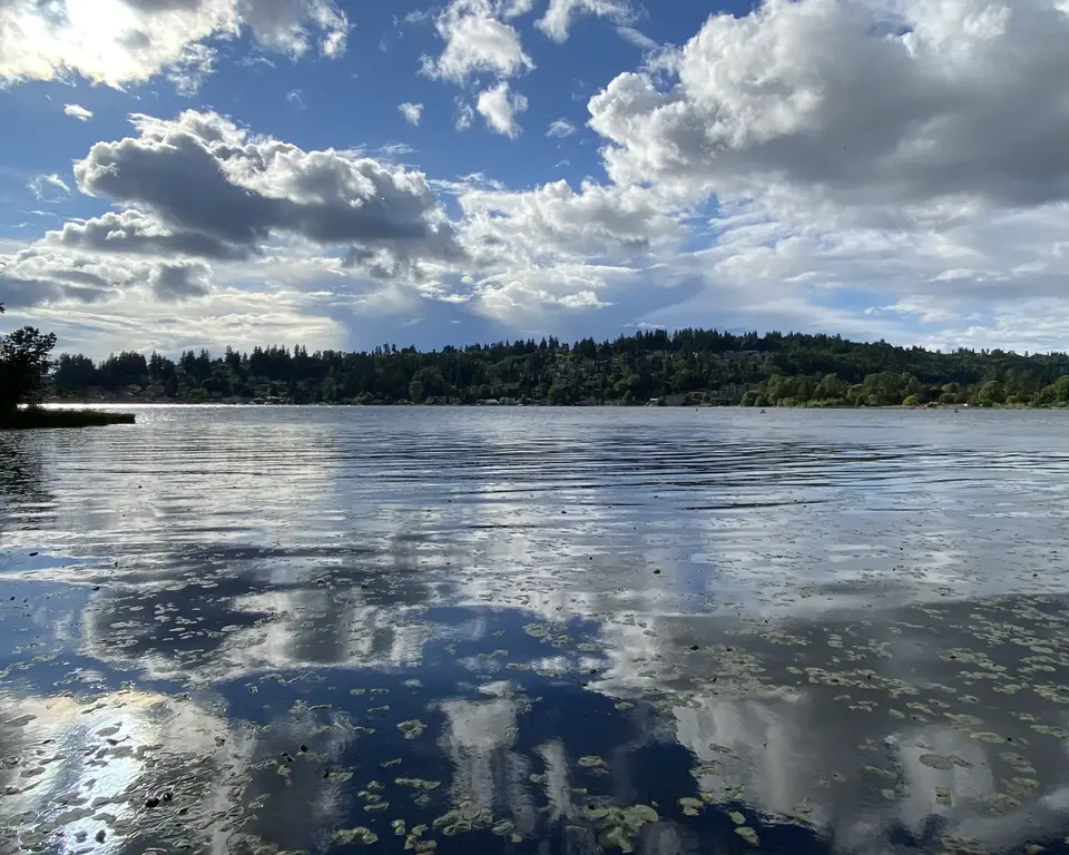 Dramatic white clouds reflect across a calm lake bordered by a forested shoreline.