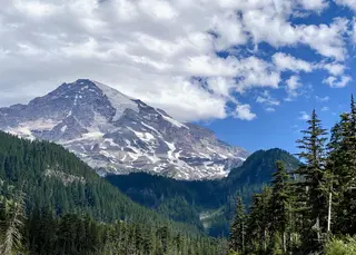 A rugged, snow-streaked mountain rises above a dense evergreen forest beneath drifting clouds and blue sky.