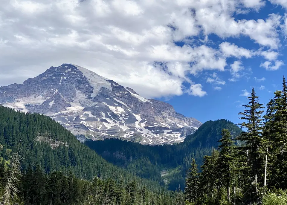 A rugged, snow-streaked mountain rises above a dense evergreen forest beneath drifting clouds and blue sky.