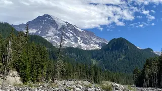 A snow-streaked rocky mountain rises above a dense evergreen forest beneath a partly cloudy blue sky.