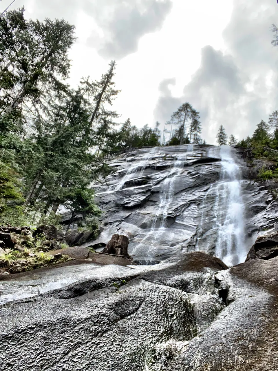 Water cascades down a broad rocky cliff surrounded by evergreen trees beneath a cloudy sky.