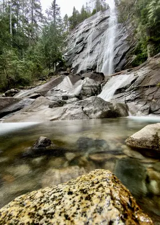 A cascading waterfall flows over dark rock slabs into a clear pool surrounded by forest.