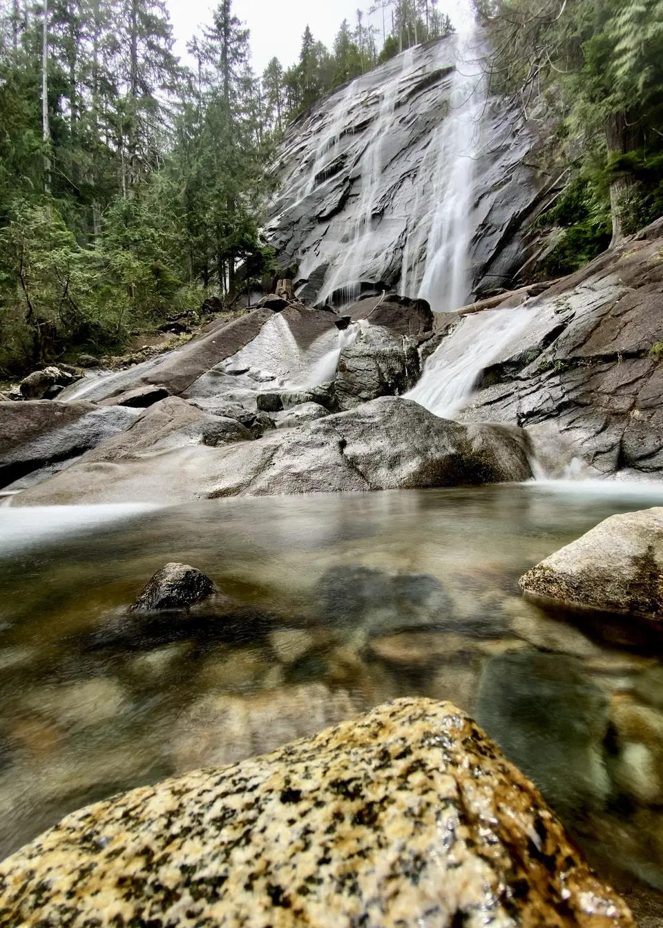 A cascading waterfall flows over dark rock slabs into a clear pool surrounded by forest.
