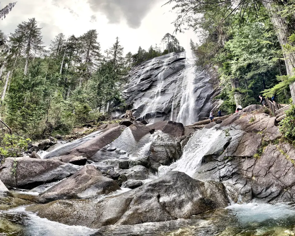 A waterfall cascades over broad dark rocks through a dense evergreen forest, with several hikers standing near the falls.