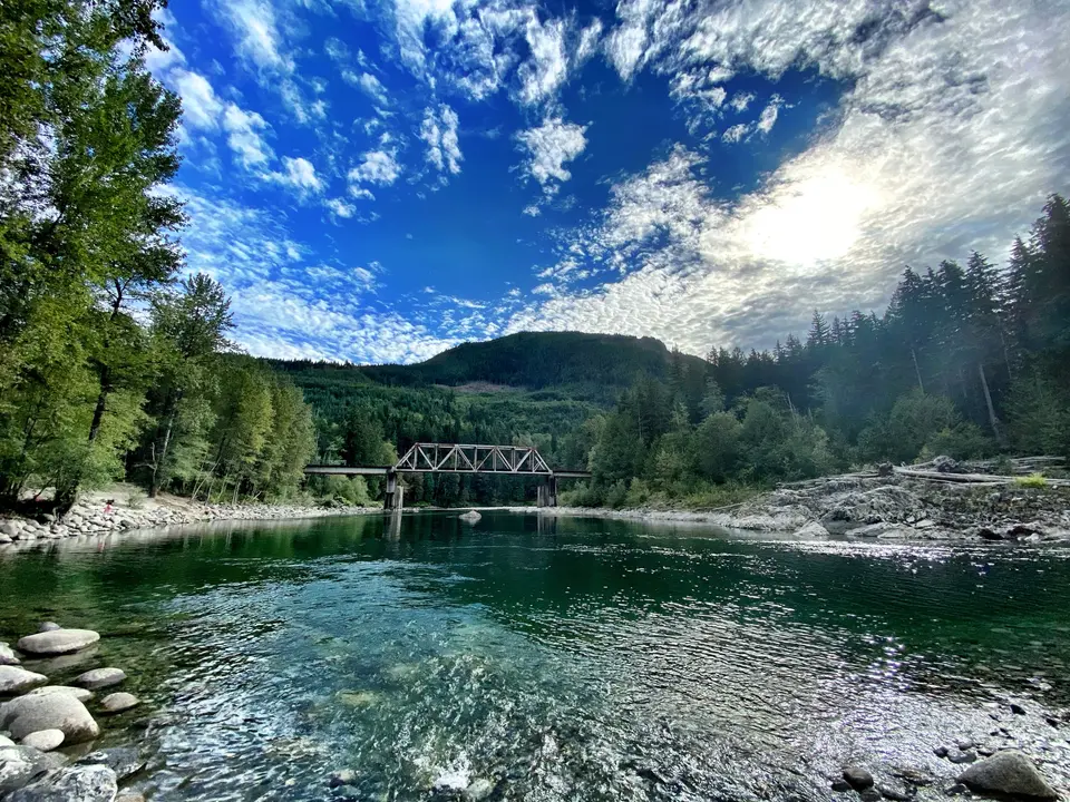 A clear river flows beneath a metal truss bridge amid a forested hillside under a dramatic blue sky.