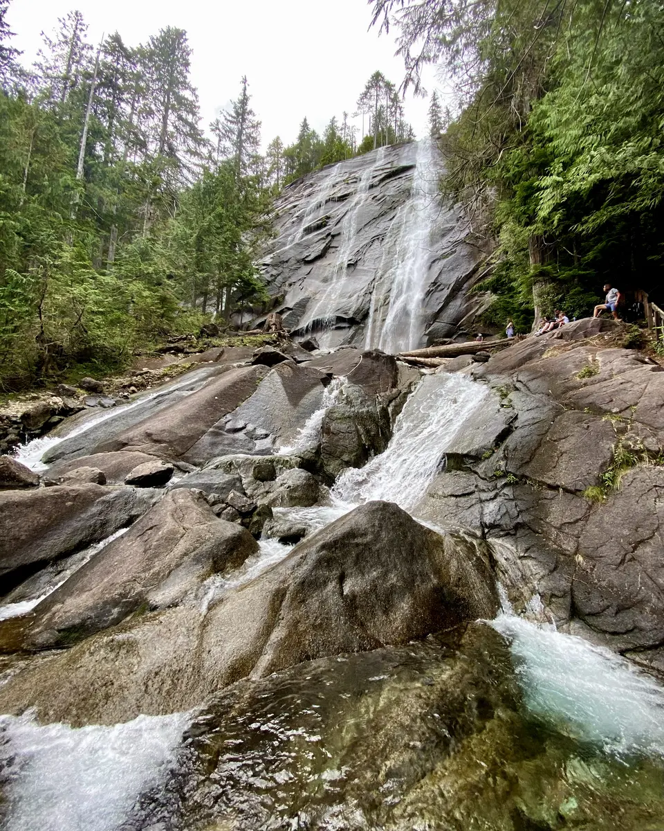 A waterfall cascades down broad dark rocks through a forest, with visitors sitting along the rocky bank.