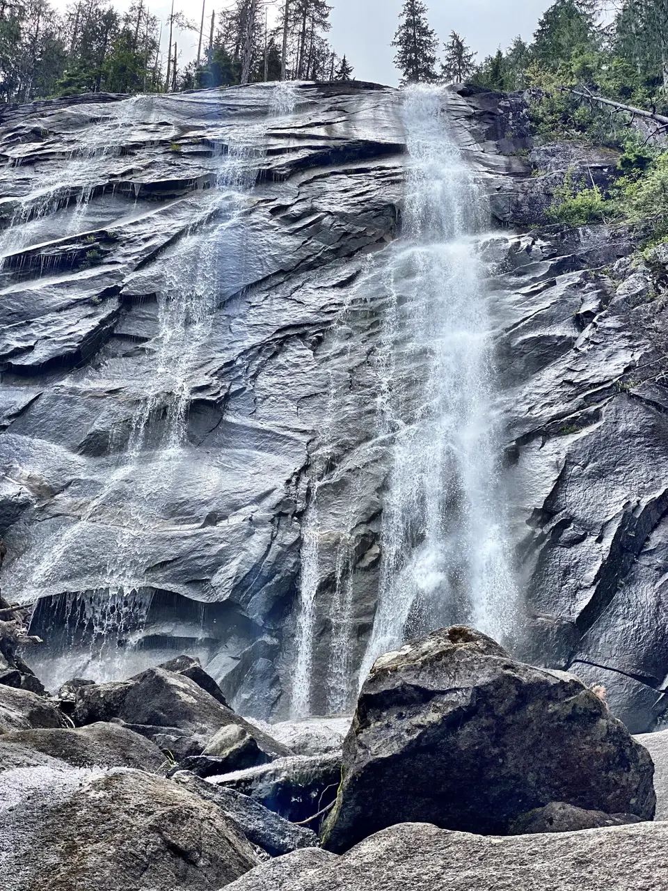 Water cascades down a broad dark rock face surrounded by evergreen trees and boulders.