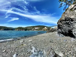 A pebbled rocky shore curves beside blue water beneath wispy clouds, with a steel bridge spanning the forested inlet in the distance.
