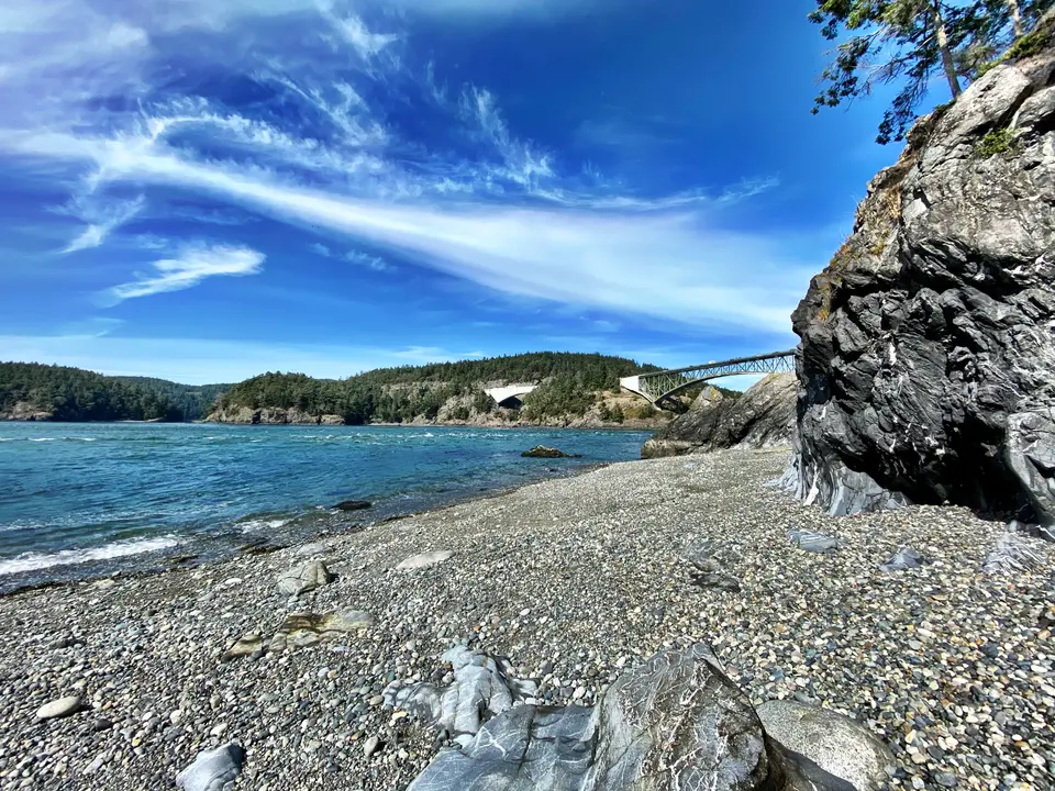 A pebbled rocky shore curves beside blue water beneath wispy clouds, with a steel bridge spanning the forested inlet in the distance.