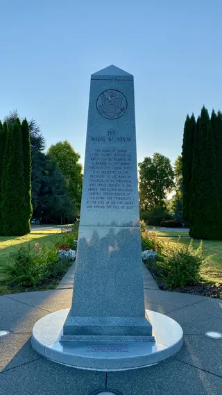A tall granite monument inscribed “The Medal of Honor” stands amid flower beds and evergreen trees beneath a clear blue sky.