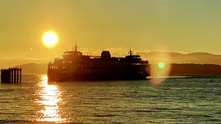 A large passenger ferry moves across rippling water in silhouette beneath a glowing sunset.