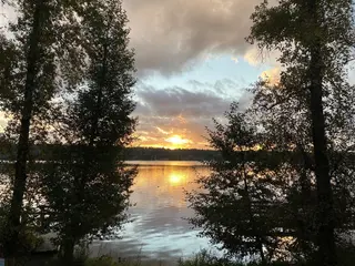 The setting sun reflects across a calm lake framed by leafy trees and a cloudy sky.