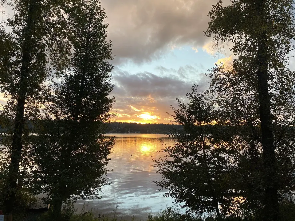 The setting sun reflects across a calm lake framed by leafy trees and a cloudy sky.