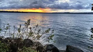 Dark clouds hang over a rippling body of water as the setting sun glows along the distant tree-lined shore.