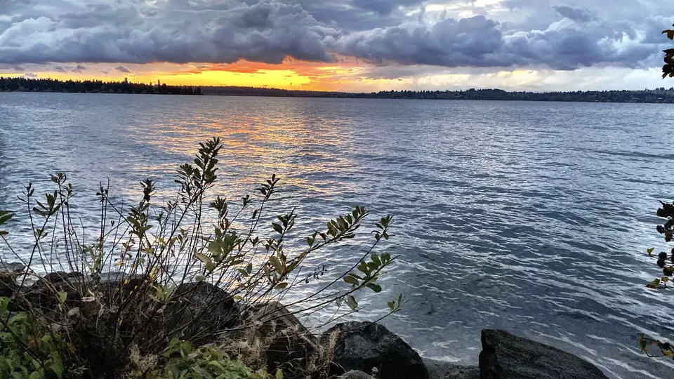 Dark clouds hang over a rippling body of water as the setting sun glows along the distant tree-lined shore.