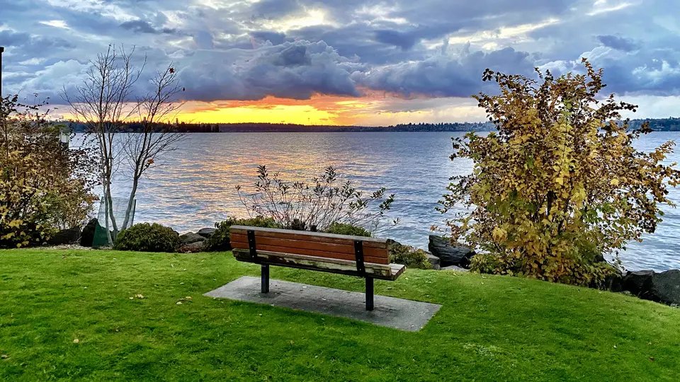 A wooden bench overlooks rippling water and autumn trees beneath a dramatic sunset sky.