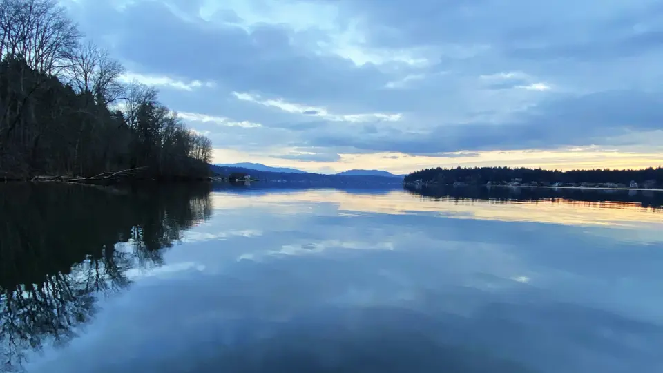 A calm lake reflects a cloudy blue sky and the dark silhouettes of trees along the shoreline at sunset.