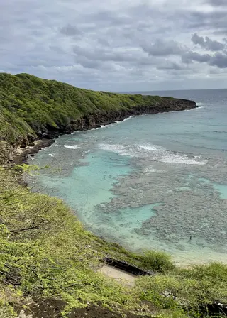 A lush rocky headland curves around clear turquoise ocean water beneath a cloudy sky.