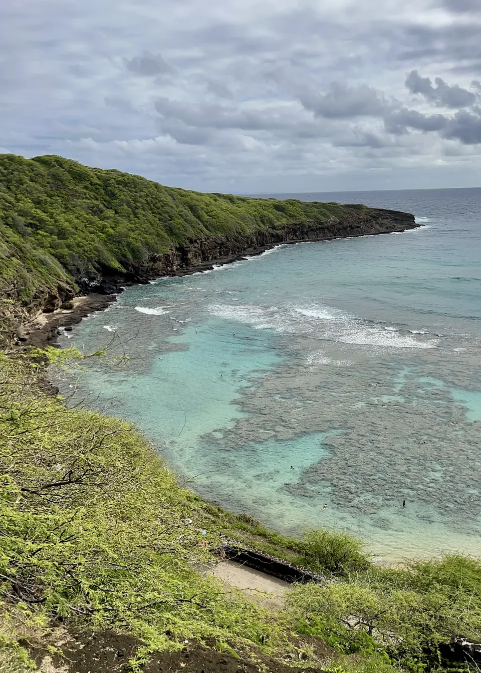 A lush rocky headland curves around clear turquoise ocean water beneath a cloudy sky.