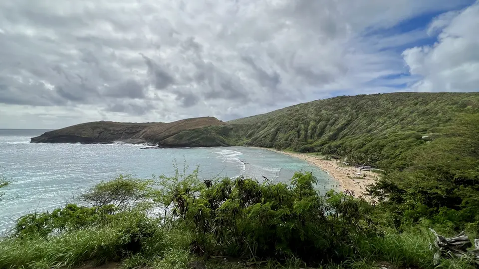 A turquoise ocean cove curves around a sandy beach beneath green coastal hills and a cloudy sky.