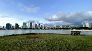 A grassy waterfront with a bench overlooks calm water and a skyline of high-rise buildings beneath scattered clouds.