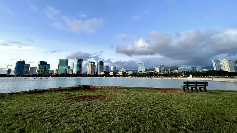 A grassy waterfront with a bench overlooks calm water and a skyline of high-rise buildings beneath scattered clouds.