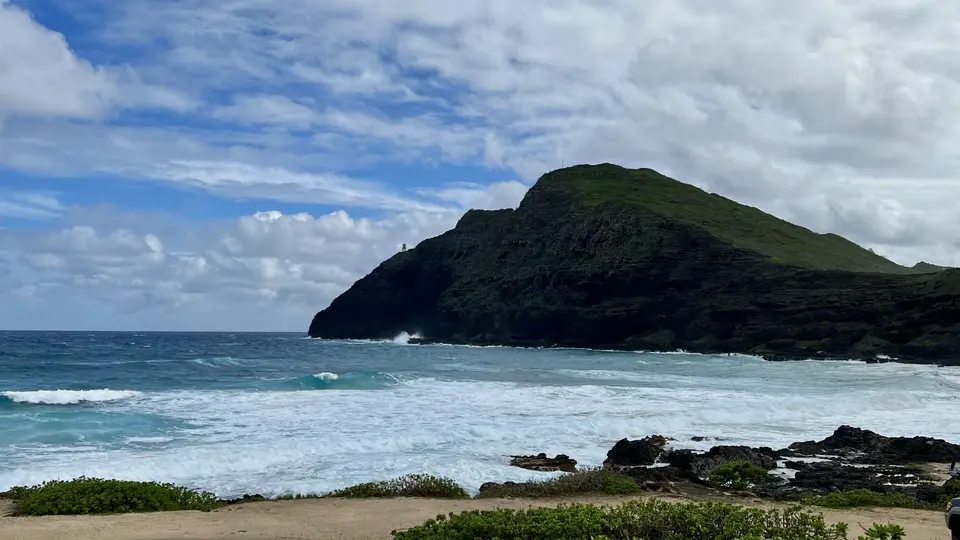 Waves roll into a rocky shoreline beneath a green coastal mountain and a cloud-filled sky.