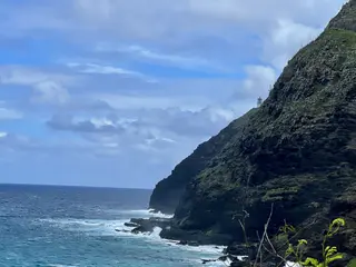 A small lighthouse stands on a steep green rocky cliff above choppy ocean waves beneath a cloudy blue sky.