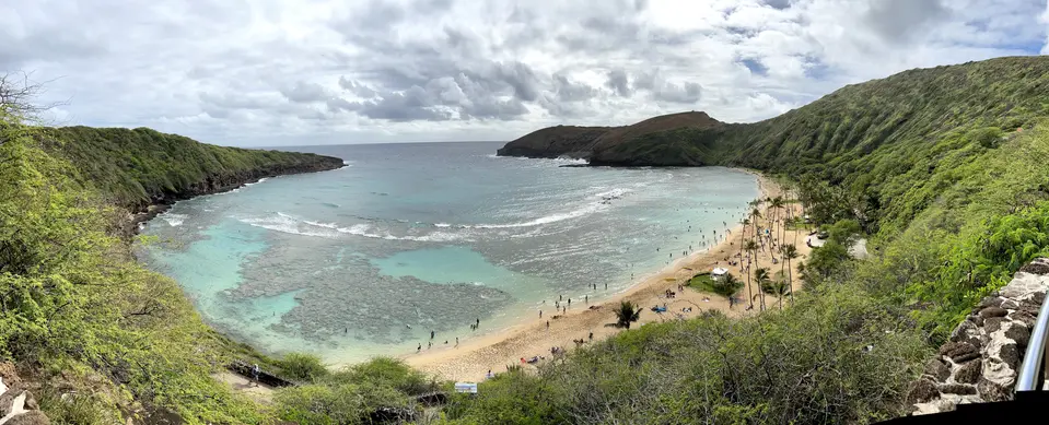 A sandy beach curves around a turquoise bay beneath lush green cliffs and a cloudy sky.