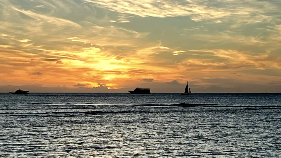 The sun sets over rippling ocean water as small boats and a sailboat silhouette the horizon.