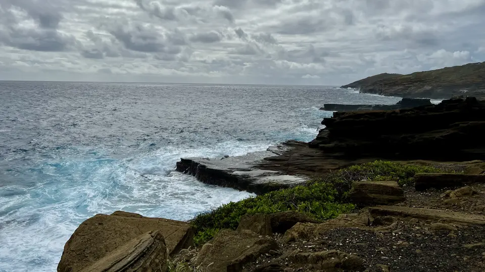 Waves crash against layered dark rock formations along a rugged ocean coastline beneath a cloudy sky.
