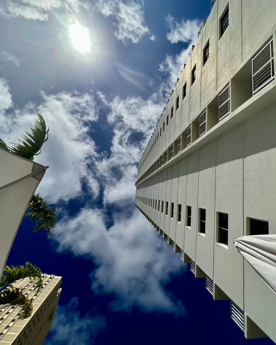 A low-angle view looks up at a white high-rise building beside palm trees beneath a bright sun and scattered clouds.