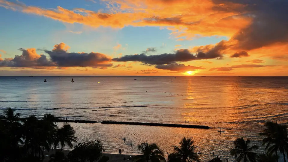 The sun sets over a calm ocean beneath vivid orange clouds, with palm trees and small boats along the shoreline.