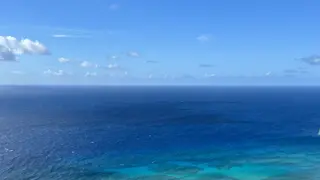 A wide expanse of deep blue ocean stretches beneath a bright sky dotted with small clouds.