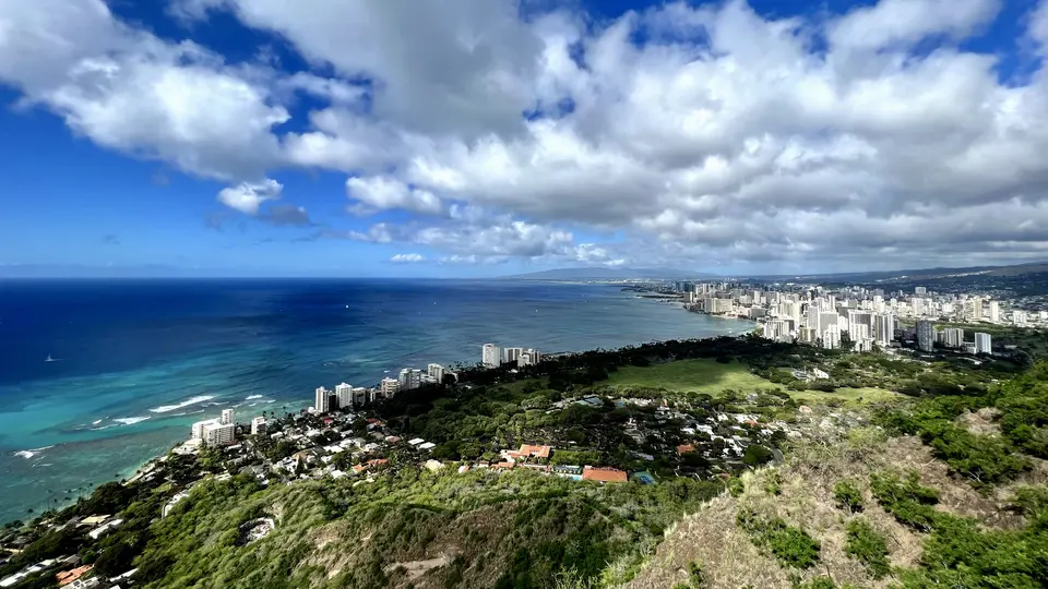 A panoramic view from a green hillside overlooks turquoise ocean water, a sandy coastline, and a dense cluster of high-rise buildings beneath dramatic clouds.