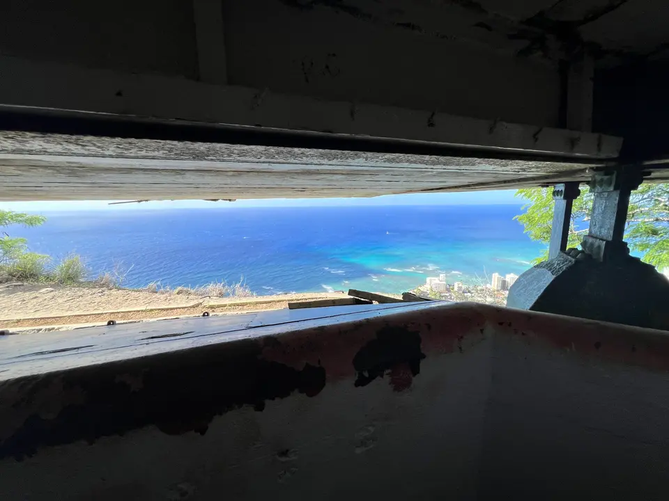 A weathered opening in a dark concrete structure frames a bright blue ocean and distant coastal buildings.