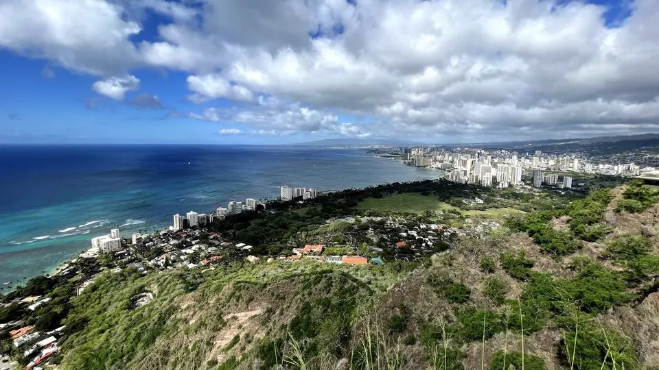 A lush hillside overlooks a turquoise ocean and a dense coastal skyline beneath broad clouds.