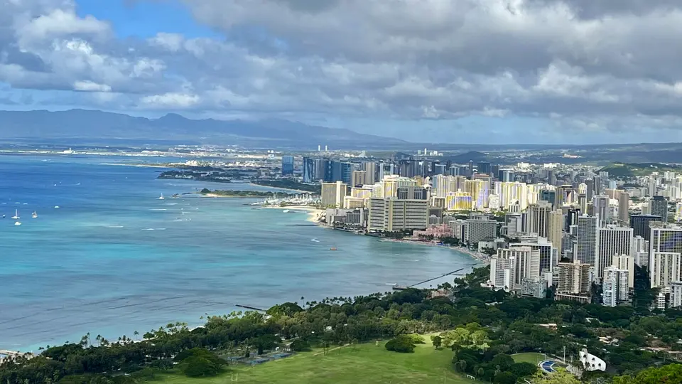 High-rise buildings line a turquoise bay beneath a cloudy sky, with green parkland and distant mountains beyond.