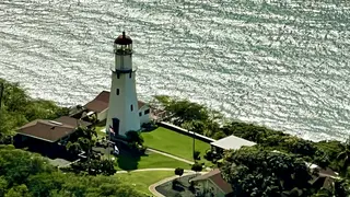 A white lighthouse with a red roof stands beside a manicured green lawn and shimmering water.