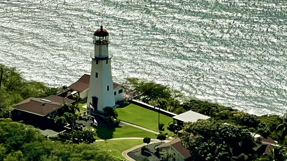 A white lighthouse with a red roof stands beside a manicured green lawn and shimmering water.