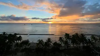 The setting sun casts an orange reflection across the ocean beyond a palm-lined beach beneath dramatic clouds.