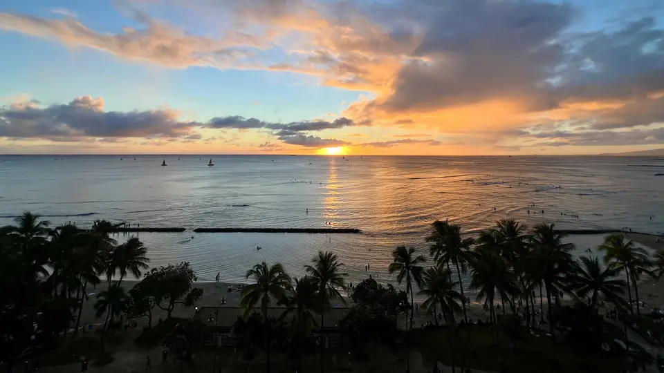 The setting sun casts an orange reflection across the ocean beyond a palm-lined beach beneath dramatic clouds.