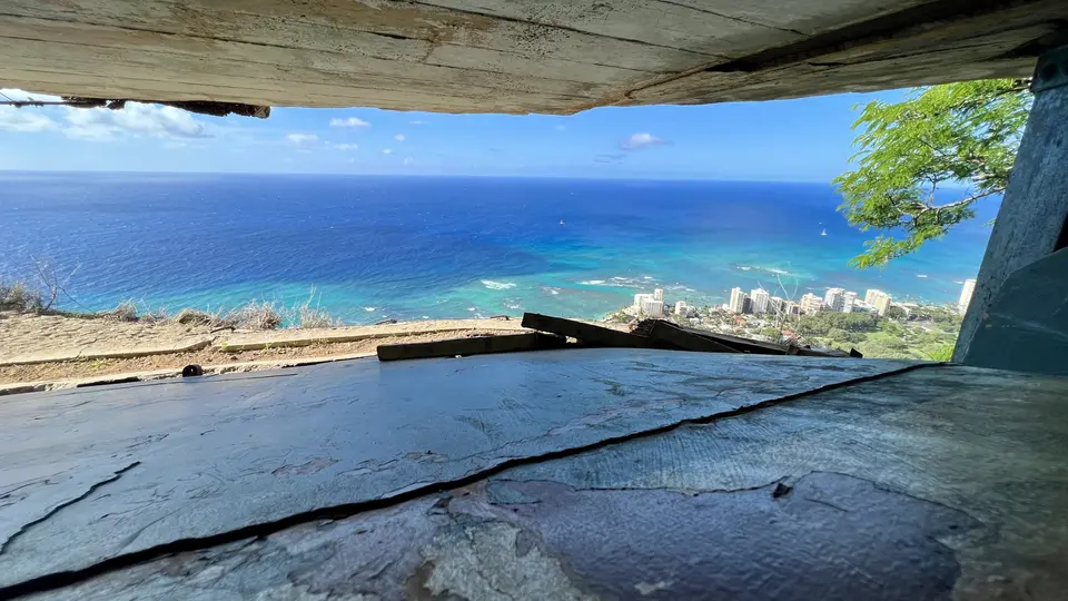 A cracked concrete floor beneath a weathered structure frames a view of turquoise ocean and coastal high-rise buildings.