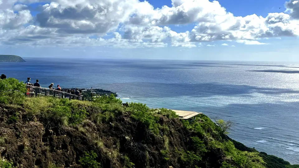 A group of visitors stands by a railing on a lush rocky cliff overlooking the ocean beneath a bright, cloud-filled sky.