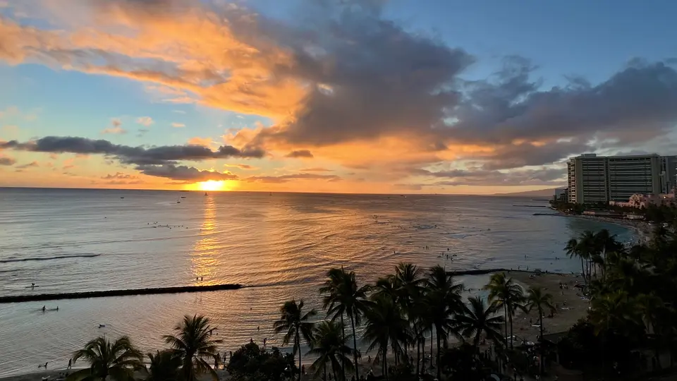 The sun sets over the ocean, casting orange reflections across the water beside a palm-lined beach and high-rise buildings.
