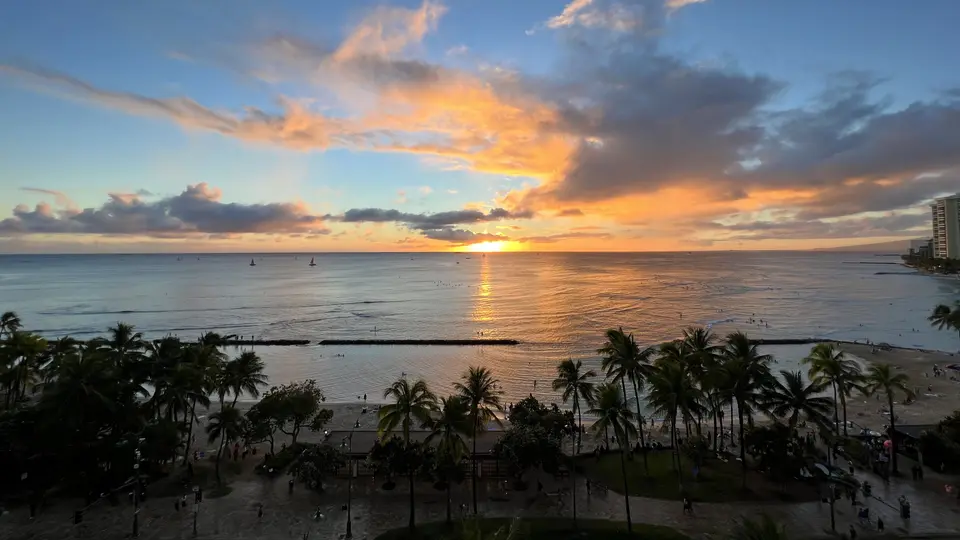 The sun sets over a calm ocean, casting a golden reflection beyond palm trees and a crowded beach.