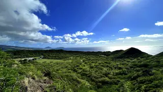 A sunlit view overlooks lush green hills and the ocean beneath a vivid blue sky filled with scattered clouds.